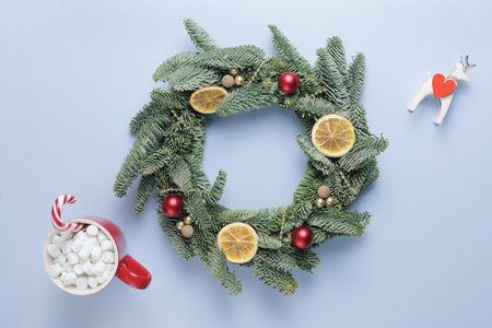 Christmas wreath made of fir branches, with small red and gold balls, slices of dried orange, shiny beads on a blue surface. A red cup of coffee with marshmallows, a candy cane and a deer figurine.の写真素材