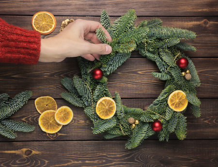 Womens hands decorate a Christmas wreath made of fir branches, with small red and gold balls, slices of dried orange, shiny beads on a wooden table. FLat lay.の写真素材