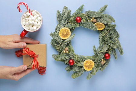Christmas wreath made of fir branches, with small red and gold balls, slices of dried orange, shiny beads and gift box on a blue surface. A red cup of coffee with marshmallows, and a candy cane. Top view. Flat lay.の写真素材