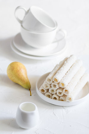 Delicate sweet cookies in the form of tubes in coconut flakes filled with vanilla cream on a white plate and a yellow pear with two tea mugs in the background.の写真素材