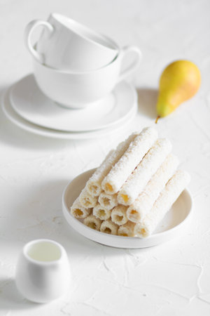 Delicate sweet cookies in the form of tubes in coconut flakes filled with vanilla cream on a white plate and a yellow pear with two tea mugs in the background.の写真素材