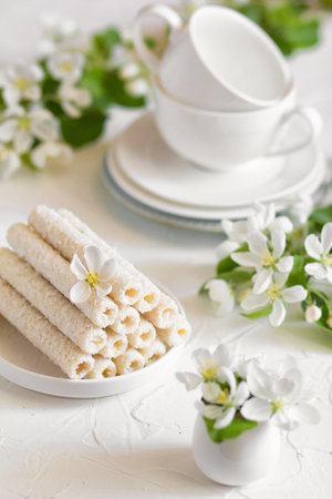 Delicate sweet cookies in the form of tubes in coconut flakes filled with vanilla cream on a white plate and a yellow pear with two tea mugs in the background.の写真素材