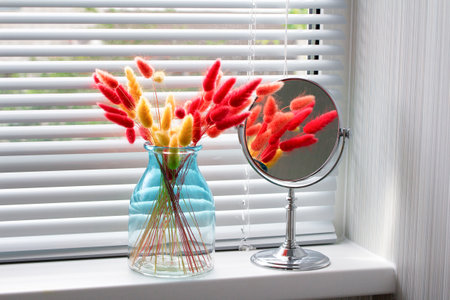 Red and yellow dry lagurus in a glass vase and a makeup mirror on the windowsill with blinds.の写真素材