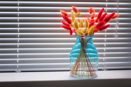 Red and yellow dry lagurus in a glass vase on the windowsill with blinds.の写真素材