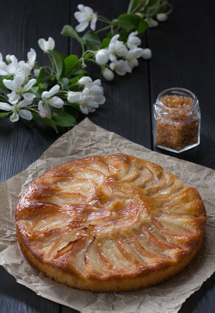 Freshly baked apple pie on parchment paper on a black table. A branch of a blooming apple tree and a can of brown sugar in the background.の写真素材