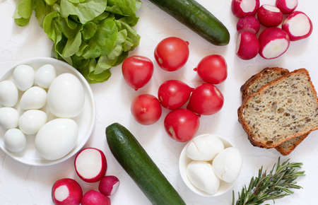 Fresh lettuce, cucumbers, tomatoes, radishes, rosemary, peeled quail and chicken eggs and mozzarella cheese on the white table. Farm products.の写真素材