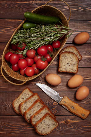 Fresh tomatoes, cucumbers, a few sprigs of rosemary in a wicker basket, chicken eggs, and sliced bread on a wooden table. Harvesting. Farm product.の写真素材