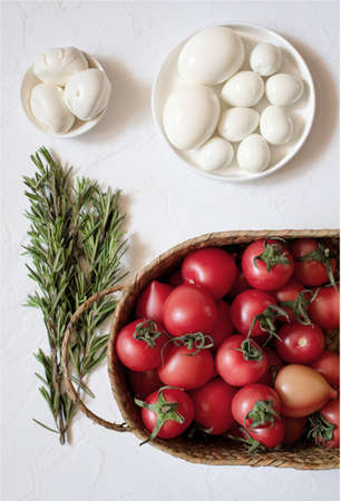 Fresh tomatoes in a wicker basket, peeled chicken and quail eggs, mozzarella cheese and a few sprigs of rosemary on a white surfaceの写真素材