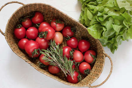 Fresh tomatoes, few sprigs of rosemary in a wicker basket, with lettuce leaves on a white surface. Harvest. Farm product.の写真素材