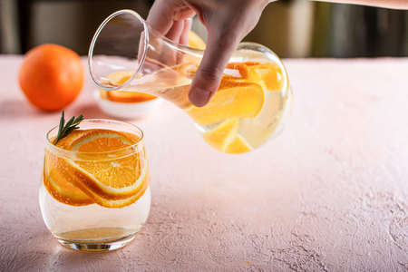 A womans hand pours a refreshing detox lemonade with sliced orange into a glass tumbler on a pink table.の写真素材