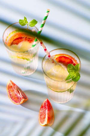 Two glasses of refreshing summer lemonade with ice, slices of lemons and red oranges with sprigs of mint on the blue table. Summer drinks. Copy spaceの写真素材