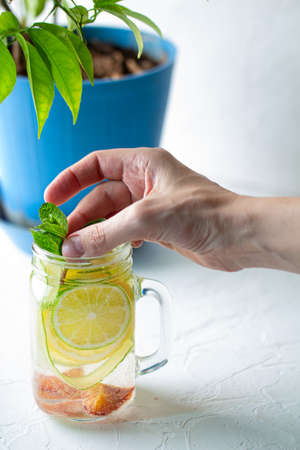 a womans hand puts down a sprig of mint into a glass mug with handle with refreshing detox lemonade with sliced lime, lemon, cucumbers and red orange on white table.の写真素材