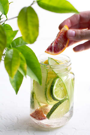 A womans hand throws a slice of red orange into a glass mug with handle with refreshing detox lemonade with sliced lime, lemon, cucumbers and red orange on white table.の写真素材