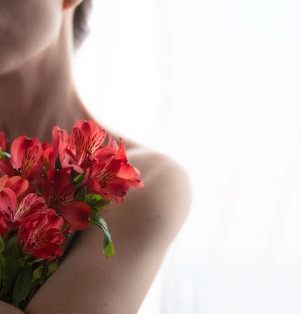 A naked girl in defocus clutches a bouquet of red alstroemeria to her shoulder on white background. Front view. Selective focus.の写真素材