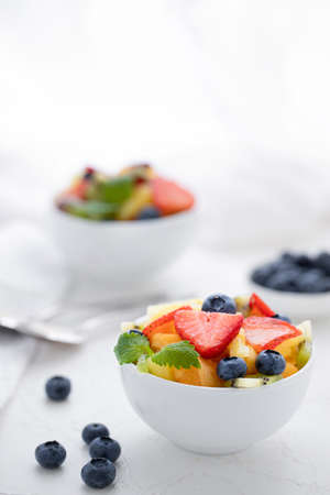 Fresh fruit salad with fresh strawberries, kiwi, apricots and blueberries, decorated with leaves of fresh mint in white plates on a white tableの写真素材