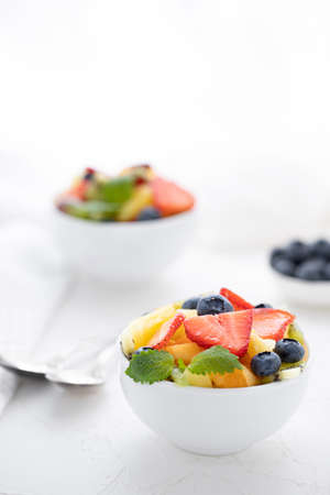 Fresh fruit salad with fresh strawberries, kiwi, apricots and blueberries, decorated with leaves of fresh mint in white plates on a white tableの写真素材