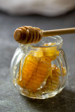 Sealed honeycomb of honey with wooden Honey Dipper for mixing and tasting natural honey in a small glass jar on a grey table.の写真素材