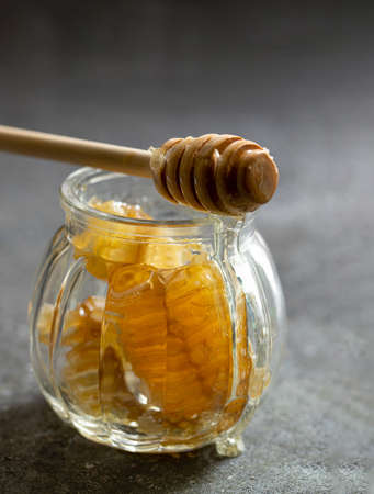 Sealed honeycomb of honey with wooden Honey Dipper for mixing and tasting natural honey in a small glass jar on a grey table.の写真素材