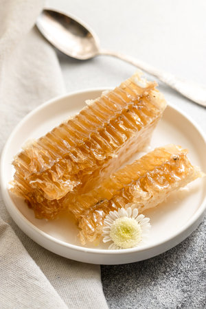 Sealed honeycomb of honey with little spoon on a white porcelain plate on a grey tableの写真素材