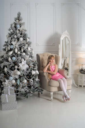 Girl in a pale pink dress sitting in a chair at the Christmas tree in a room with white interior.の写真素材