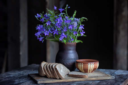 A rustic still life with a bunch of bluebells in a ceramic vase and a bowl of sliced bread on a board.の写真素材