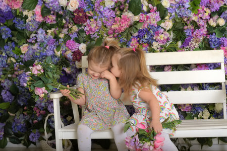 two little twin girls in the garden on a bench against the background of a floral wall with bouquets of flowers in their handsの写真素材