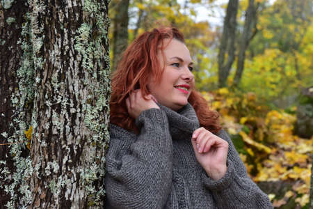 red-haired woman in a gray sweater in the autumn near a tree covered with mossの写真素材