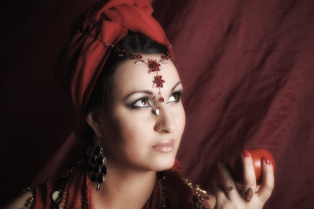 beautiful brunette woman in red oriental clothes with a turban and jewelry on her head and an apple...