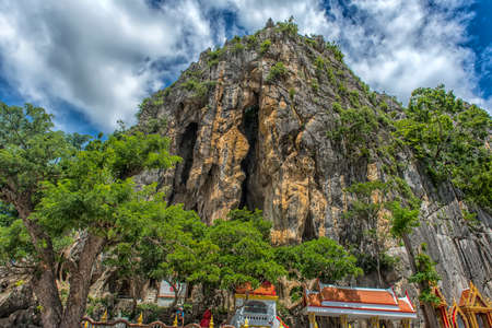 Thailand, Phetchaburi 19.07.2019 view of Wat Tham Khao Yoi Cave, Buddhist temple with green tree and rock mountain background, in Phetchaburi, Thailand with Thai text banner means the name of this local is Wat Tham Chao Yoi Cave.のeditorial素材