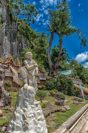 Thailand, Phetchaburi 19.07.2019 view of Wat Tham Khao Yoi Cave, Buddhist temple with green tree and rock mountain background, in Phetchaburi, Thailand with Thai text banner means the name of this local is Wat Tham Chao Yoi Cave.のeditorial素材