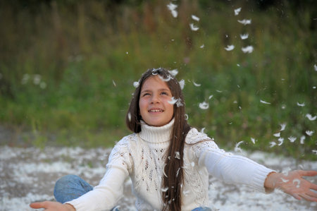 young girl with long hair in a white sweater plays happy among white feathers