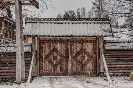 massive wooden gates decorated with intricate carvings in traditional Russian style. The gates are part of a fortified wooden structure, probably a fort or a medieval manor. The wood looks aged, and the ornaments give the structure an authentic look. The roof above the gates is covered with a layer of snow, which emphasizes the winter atmosphere. Snow-covered trees grow around, and the road in front of the gates is strewn with snow, in places covered with footprints. The whole scene conveys the spirit of ancient Rus', village life and historical architecture.の写真素材