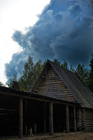 a traditional wooden house located in the middle of the forest. The house has a gable roof with deer antlers hanging from it, which may indicate its connection with hunting traditions or the symbolism of ancient peoples. The faÃ§ade is made of logs, and a canopy is attached to the house, supported by massive wooden posts.

Dramatic, massive thunderclouds rise in the background, creating a contrast between the calm village architecture and the approaching storm. Light breaks through the cloud layer, giving the scene a mystical and even menacing look. Dark shades enhance the atmosphere of mystery and antiquity.の写真素材