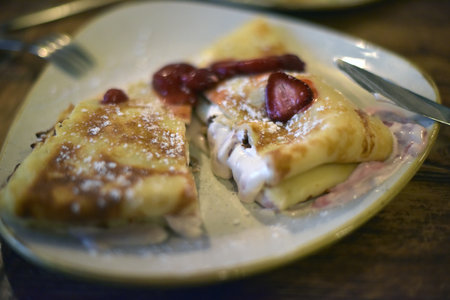 pancakes with a filling of butter cream and strawberries, served on a plate. The pancakes have a golden crust and are sprinkled with powdered sugar. The filling flows out slightly, creating an appetizing effect. Strawberry pieces on top give the dessert freshness and a bright color accent. The cozy atmosphere of a cafe or home feast is visible, and in the frame there are cutlery - a knife and fork, ready for a meal.の写真素材