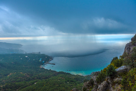 stunning view of the Crimean coast from above. In the foreground you can see a rocky area covered with dense green vegetation, smoothly descending to the shore of the Black Sea. In the background are small buildings and residential complexes located along the coastal zone.

The most impressive detail of the photo is the rain front hanging over the sea. The boundary between the clear and rainy zone is clearly visible, where streams of rain descend from the clouds and create the effect of a blurred horizon. This spectacle emphasizes the unique microclimate of Crimea, where mountains, sea and clouds form unpredictable weather phenomena.の写真素材