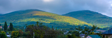 picturesque Crimean mountains covered with dense forest. Summer landscape turns into autumn, with yellowed areas of vegetation. Thick clouds envelop the mountain peaks, creating an atmosphere of fog and mystery. Houses are visible below, the roofs of which are slightly covered by the greenery of the trees. In the distance, you can see individual buildings and roads leading to mountain villages. The nature of Crimea in this picture looks peaceful and inspiring.の写真素材