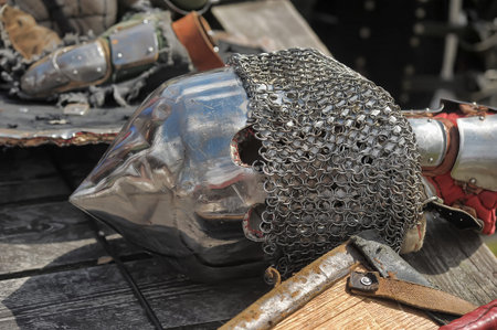 medieval knight's helmet - bascinet with aventail (chainmail protection for the neck). The helmet is made of metal, polished to a shine, but has traces of scratches and abrasions, which indicates its active use, possibly in historical reconstructions or buhurts. Breathing holes are visible, as well as chainmail protecting the warrior's neck.

Nearby are elements of armor: gauntlets, parts of bracers and fragments of a shield. The helmet with a pointed shape was popular among the knights of Europe in the 14th-15th centuries and was used in battle along with a cuirass and plate armor.の写真素材