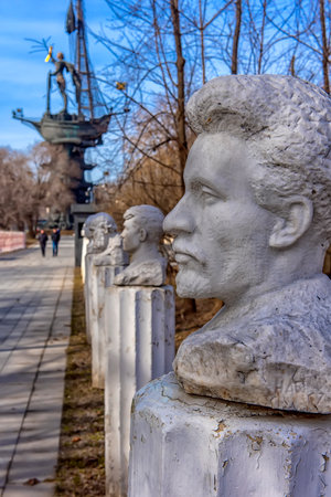 The photo was taken in the "Arts Park Museum" in Moscow, known as the "Museum Park" or "Fallen Idols Museum." The frame shows an alley of busts, among which are probably monuments to Soviet figures, removed from their pedestals and moved here after the collapse of the USSR.

In the foreground, a plaster or concrete bust of a man with wavy hair and a characteristic beard is clearly visible. Along the path are other busts standing on cylindrical pedestals.

In the background, the monument to Peter the Great, created by Zurab Tsereteli, rises. This monument is one of the most famous and controversial in Moscow, as many consider it excessively massive and disproportionate.

The lighting and dry trees indicate that the photo was taken in early spring or late autumn.の写真素材