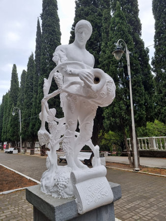 a snow-white sculpture depicting an ancient youth with an amphora and a grapevine. This is a classic allegory of fertility and abundance, executed in an idealized style. At the foot of the statue, the word is engraved on a scroll, indicating the theme of winemaking. The sculpture stands on the street in the city of Simeiz, against the backdrop of a cypress alley and park benches, creating the atmosphere of a southern resort with notes of ancient aesthetics and Crimean hospitality.の写真素材