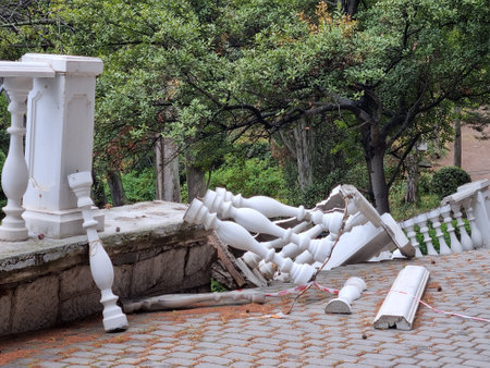 a collapsed section of a balustrade, made in the classical style with white balusters. It has partially collapsed down from a terrace or path framed by stonework. The debris lies on a paved path, part of the fence is still in place. It is clear that the area was fenced off with red and white tape for safety. There is green vegetation, trees and bushes around. The place looks like a park or embankment, perhaps this is Simeiz, Crimea.の写真素材