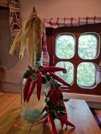 a cozy corner of a traditional rural house. On a wooden table there is a glass bottle with an ear of corn with dried leaves stuck into it, and a branch with bunches of red and green capsicum is entwined around the neck. In the background there is a window with a wooden frame and curtains decorated with red folk embroidery. Green tree branches are visible through the window glass, creating an atmosphere of coziness, coolness and rural tranquility. The interior is filled with the spirit of folk traditions, natural materials and symbols of harvest, protection and well-being.の写真素材