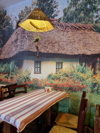 a cozy corner in the interior of a cafe or restaurant, decorated in a traditional village style. The central place is occupied by a wooden table covered with a fabric with a folk ornament. A wicker lampshade with a soft warm light hangs above the table. On the wall there is a large fresco or photo panel depicting a Ukrainian or southern Russian hut with a thatched roof, surrounded by blooming garden flowers: red, yellow and orange. The atmosphere is warm, soulful and conducive to a quiet rest.の写真素材