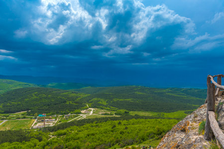 a breathtaking view from a mountain ledge in the Chelter-Marmara area of ââCrimea. In the foreground is the edge of a cliff with a wooden fence, reminding us of the presence of a hiking trail and an observation deck. In the distance are endless green spaces covered with dense forests and smoothly turning into hills. The dramatic sky with heavy blue clouds creates a sense of the grandeur of nature, its power and mystery.

This view seems to invite reflection, inspire and return to the inner silence that is so valuable in places like Chelter-Marmara - an ancient monastery hidden in the rocks.の写真素材
