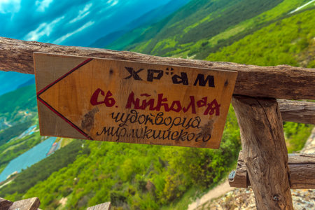 a wooden sign attached to the railing, against the backdrop of a Crimean mountain landscape. The inscription on the sign reads: "The Church of St. Nicholas the Wonderworker of Myra" - this is a landmark to the main cave church of the Chelter-Marmara complex. The sign is made by hand: a combination of black, red and handwritten letters emphasizes its warmth and organicity in the natural surroundings. The view behind the sign reveals green hills and a valley flooded with light, which makes the frame especially lively and symbolic.の写真素材