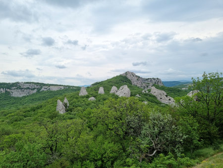 picturesque natural landscape - green hills of Crimea, rising above the forest, among which unusual rock formations protrude. This is a mysterious and mystical place near Mount Ilyas-Kaya. Stone blocks, rising above the greenery, seem to be arranged in a circle, like ancient megaliths, which gives the landscape a mysterious feel. The cloudy sky complements the atmosphere of an ancient sanctuary, preserving the silent power of nature and time.の写真素材