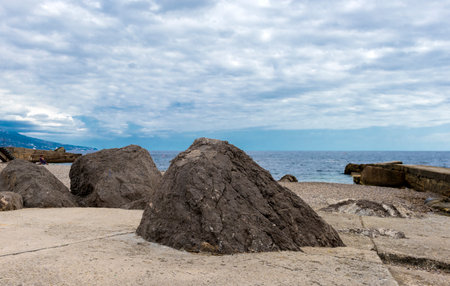 The coastline in Foros (Crimea) with massive coastal boulders in the foreground. The stones are located on a concrete platform by the sea, with pebbles and a breakwater visible nearby. Behind the calm sea surface, the horizon is hidden under a thick layer of clouds, creating an atmosphere of peace and slight drama. On the left side of the frame is the coastline, stretching into the distance, with silhouettes of mountains and buildings. Nature, minimalism and monumentality are combined in this seascape.の写真素材