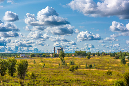 a rural or suburban landscape on a clear day. In the center of the frame rises an industrial building, perhaps a grain silo or factory, surrounded by low buildings. A spacious field with sparse trees stretches in the foreground. The main emphasis is on the sky, which takes up most of the frame and is dotted with beautiful cumulus clouds, creating depth and an aerial perspective. The lighting is soft, with daytime contrast, emphasizing the terrain and the volume of the clouds.の写真素材