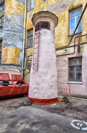 a typical courtyard of St. Petersburg with characteristic architecture and traces of time on the facades of the buildings. In the center of the frame is a ventilation shaft with a metal staircase, reminiscent of a turret. Nearby are garbage containers and several street cats that have gathered at the base of the structure. The facades of the houses are peeling and covered with stains, which emphasizes the atmosphere of an old St. Petersburg courtyard. The scene is permeated with the spirit of everyday life, comfort and city life behind the scenes of tourist routes.の写真素材
