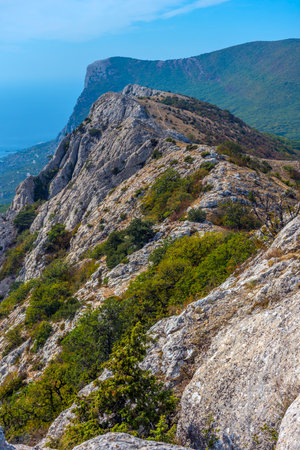 Mountain range in Crimea with picturesque cliffs covered with bushes and low-growing trees. The shooting is done from a height, opening a panoramic view of the mountain slopes going down to the Black Sea. In the background are forested hills, in the distance you can see the coastline. Bright sun, blue sky and clean air create an atmosphere of wild nature and peace.の写真素材