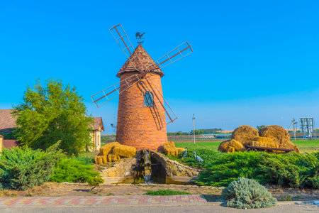 a decorative brick mill with metal blades, installed in a well-kept area against the backdrop of a clear blue sky. At the foot of the mill there is a small decorative waterfall with stonework, on the sides there are bales of hay, and nearby there are decorative figures of storks. In the background you can see green fields, some buildings and outbuildings. The whole scene looks like part of a themed recreation area in the style of village aesthetics - everything is well-kept, neat and atmospheric.の写真素材